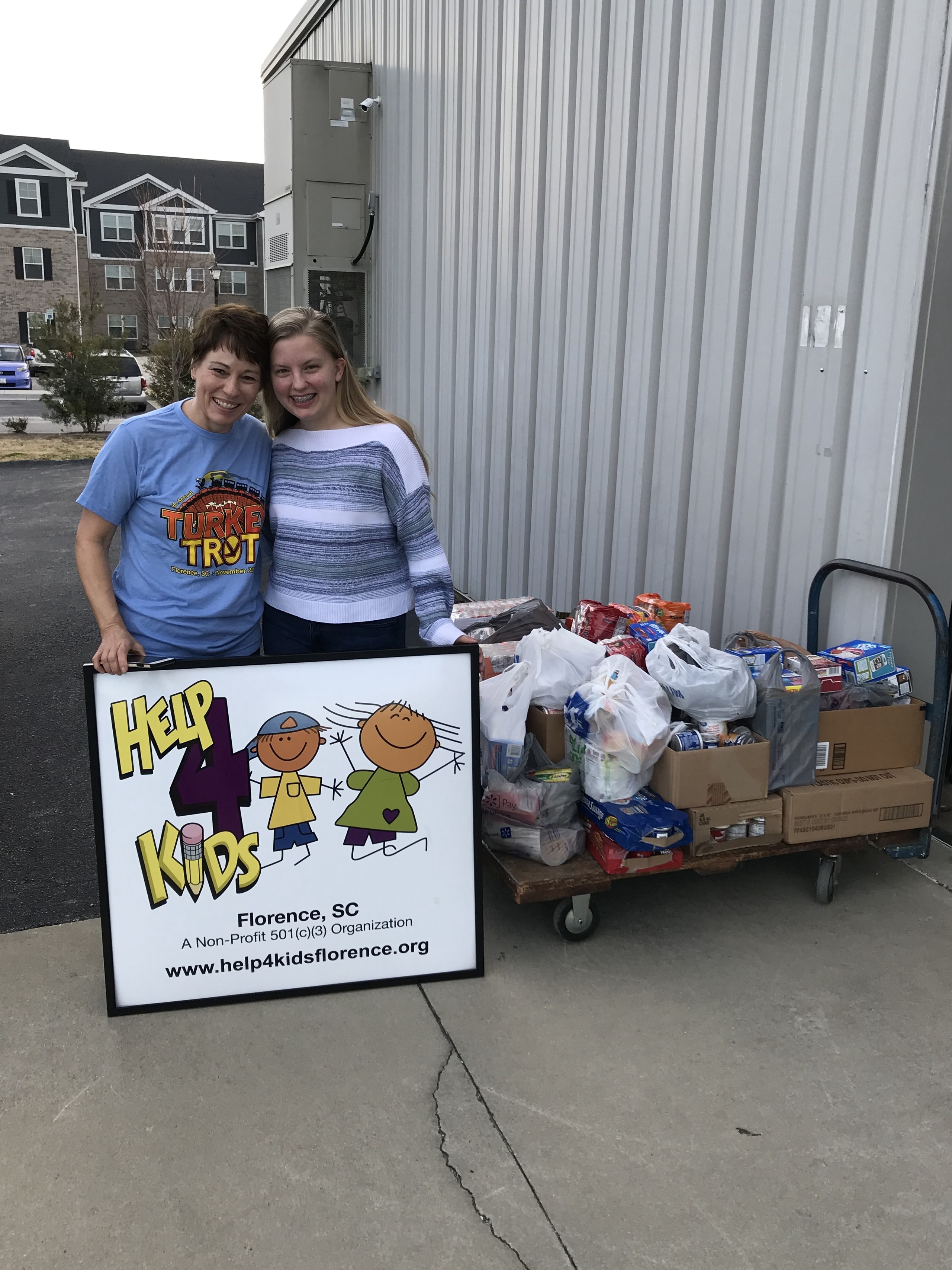 Two individuals smiling while holding a sign for Help 4 Kids, a nonprofit organization in Florence, SC. Behind them, a cart filled with donated food items for community service efforts is visible.