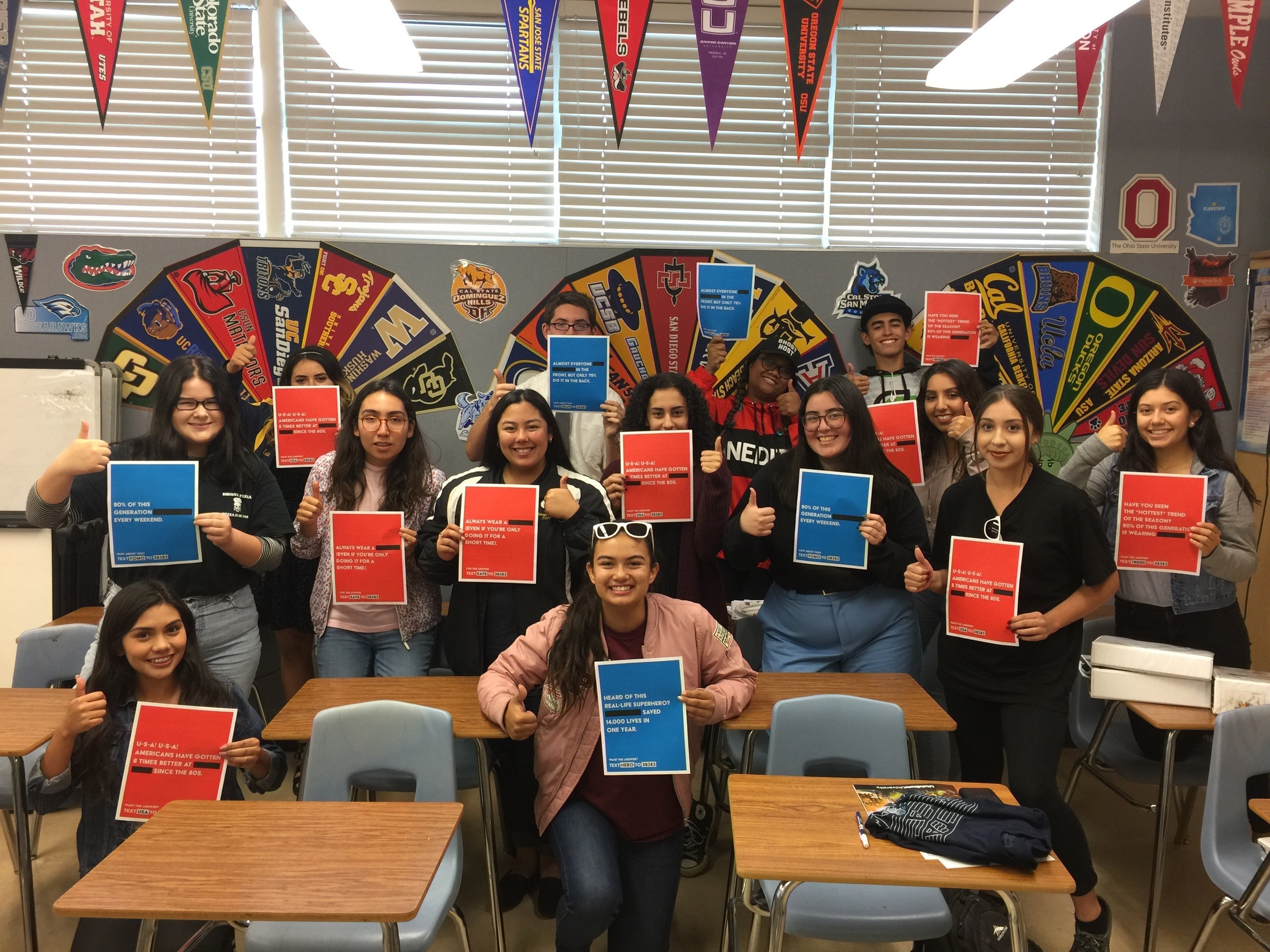 A diverse group of individuals stands in a classroom, all smiling and giving thumbs up while holding colorful flyers promoting volunteer opportunities and civic engagement. The classroom is decorated with various college pennants, highlighting an environment focused on education and community service.