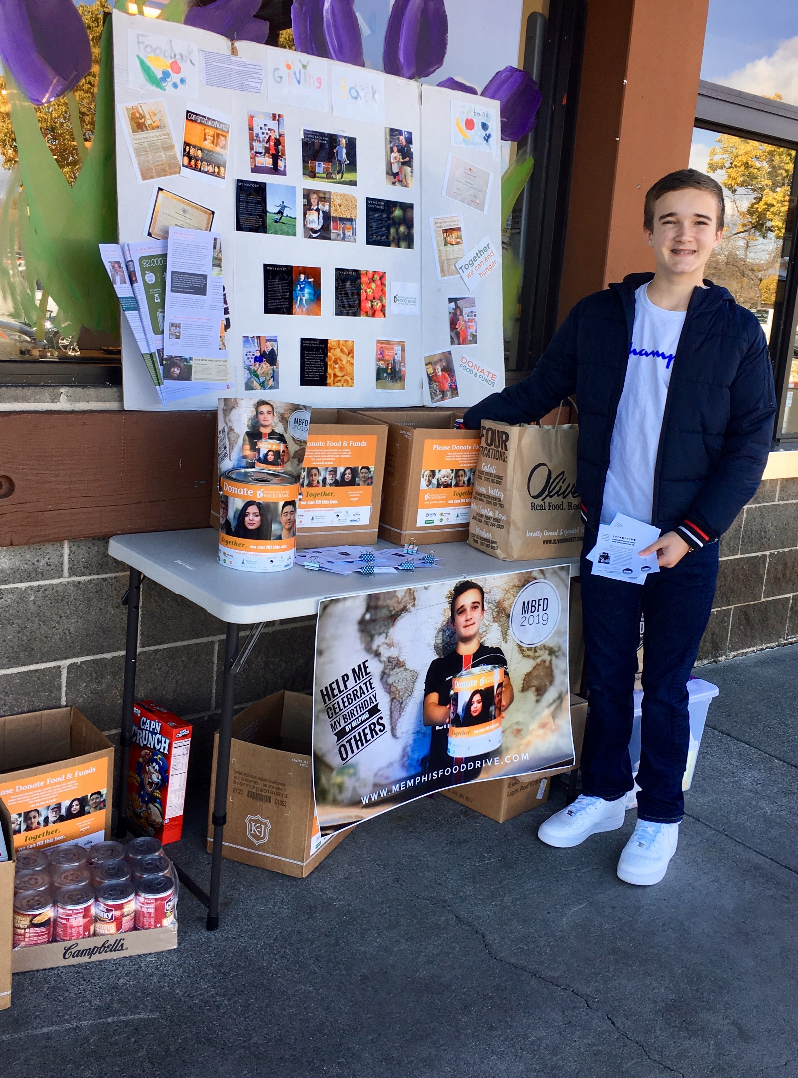 A person stands next to a table displaying volunteer opportunities for community service, including donation boxes and informative posters. The backdrop showcases community initiatives, promoting civic engagement and support for local food drives.