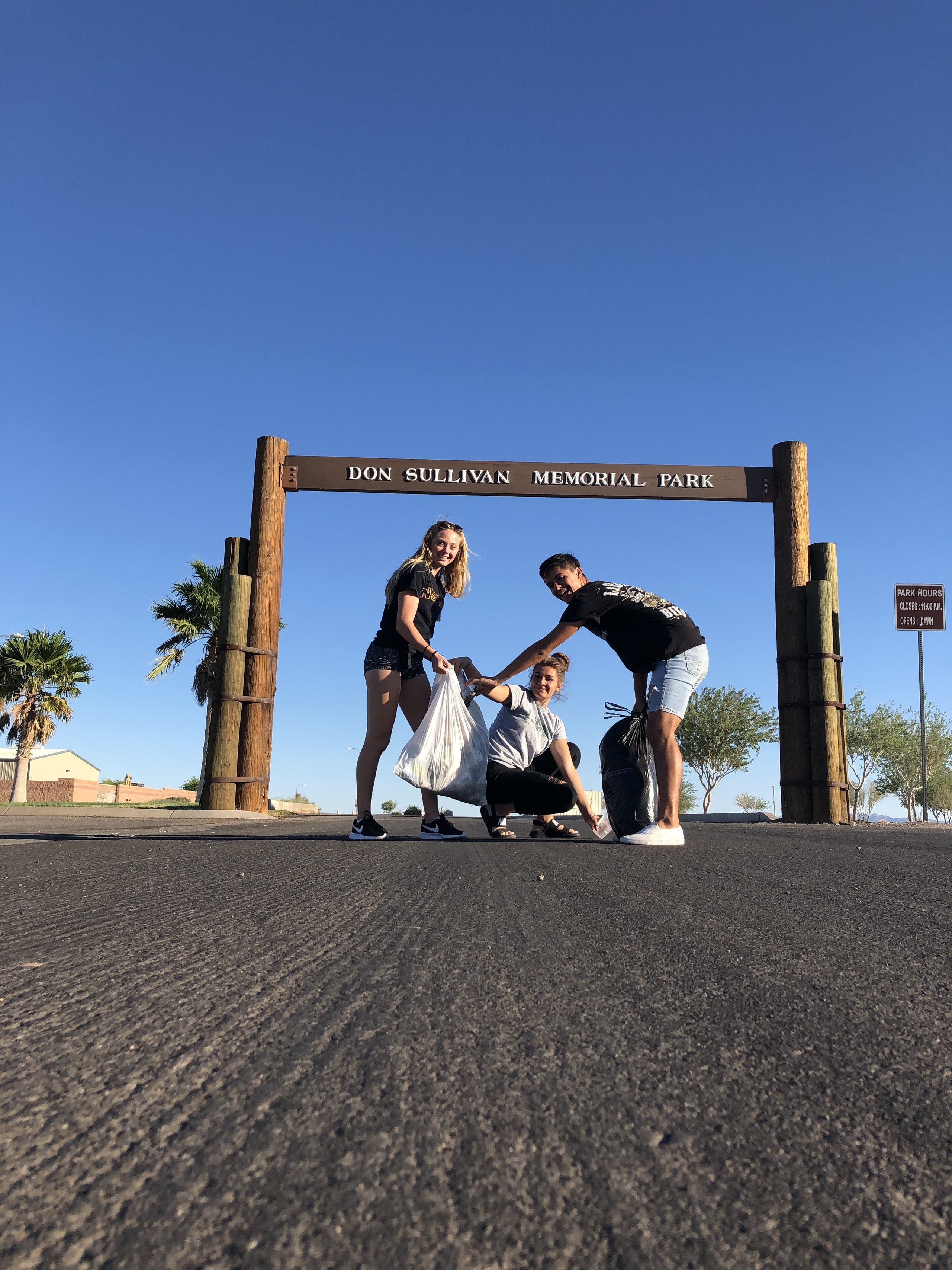 Three individuals participate in a community clean-up at Don Sullivan Memorial Park, holding trash bags while smiling under a clear blue sky. This image promotes volunteer opportunities and civic engagement in environmental stewardship.