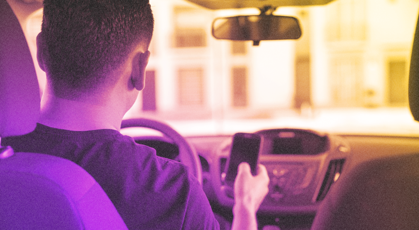 A young person with close cut hair driving a car while also using a cellphone. The vantage point is from the backseat passenger side.