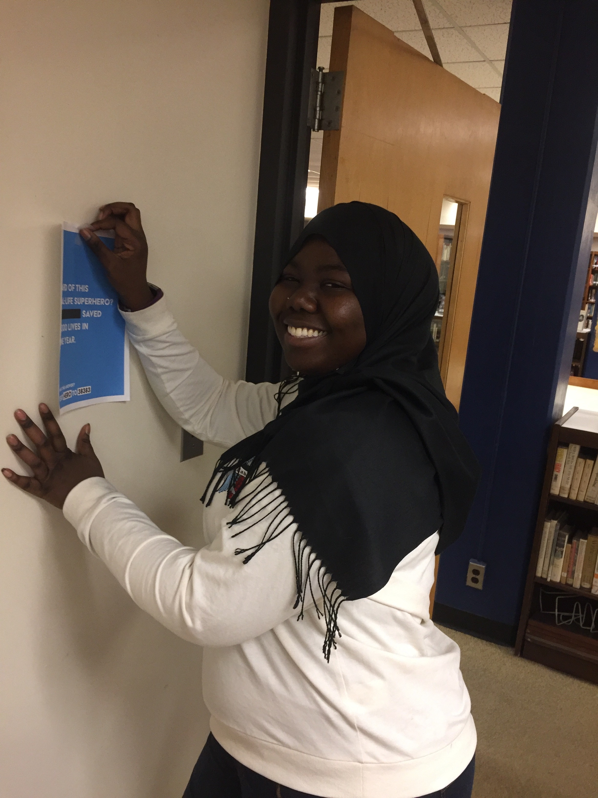 A smiling individual wearing a white sweatshirt and a black scarf posters a flyer on a wall in a library. The flyer promotes community service opportunities and emphasizes the impact of volunteering.