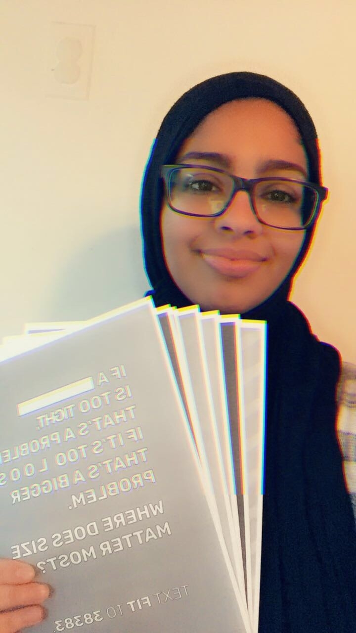 A person wearing a hijab and glasses smiles while holding several informational flyers that promote community service opportunities and civic engagement. The flyers feature bold text discussing issues related to social justice and community involvement.