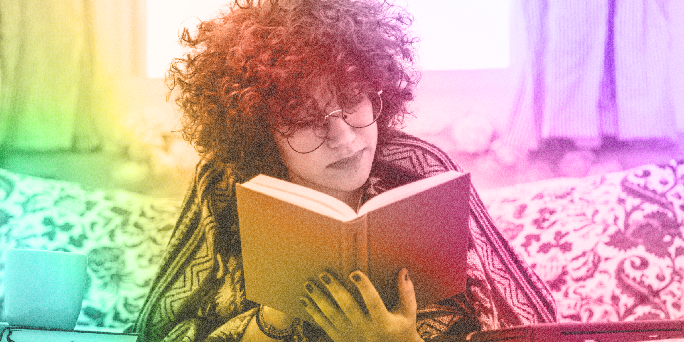 Young person with puffy curly hair and glasses reading a book while seated on a patterned couch