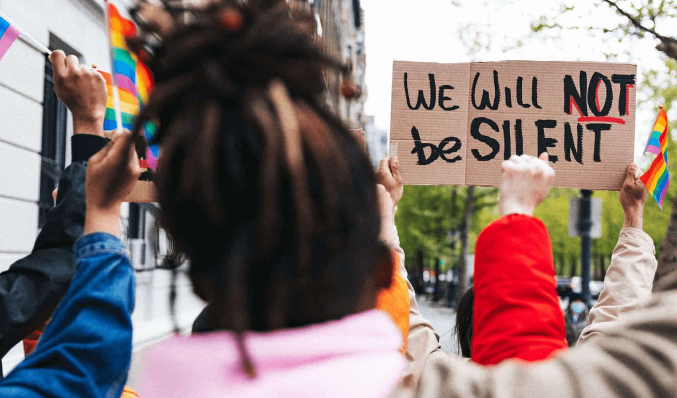 Young activists exercise their civic voices in a peaceful protest; a cardboard sign reads ' We will NOT be SILENT', while others wave rainbow pride flags.