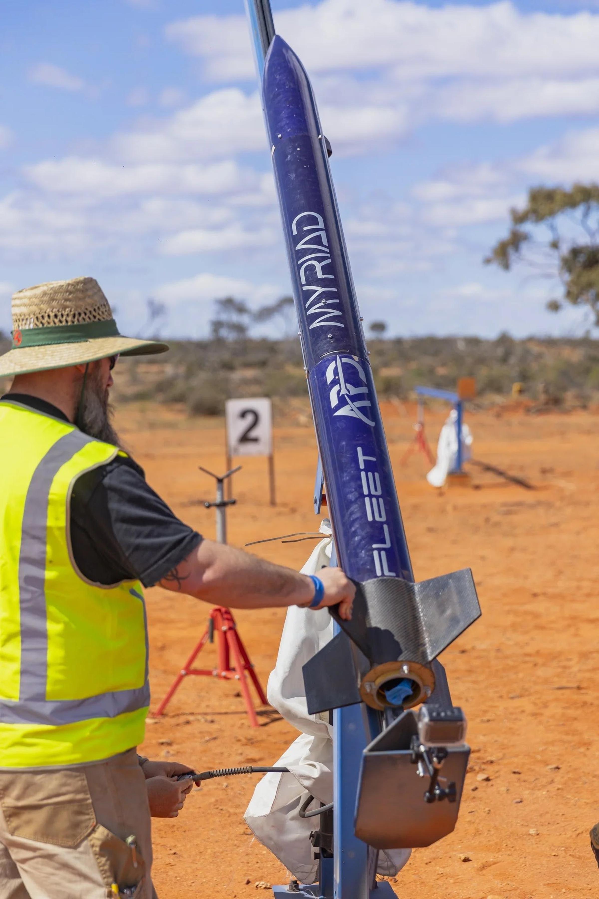 A range official in a hi-vis vest and wide-brimmed hat works at the base of the navy Myriad rocket on a blue launch rail.