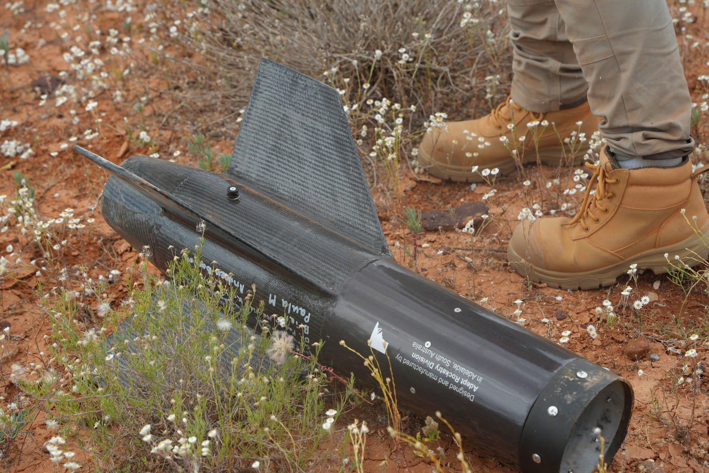 The black aft section of Kronos with swept black fins lies on red soil among white wildflowers, next to a pair of work boots.