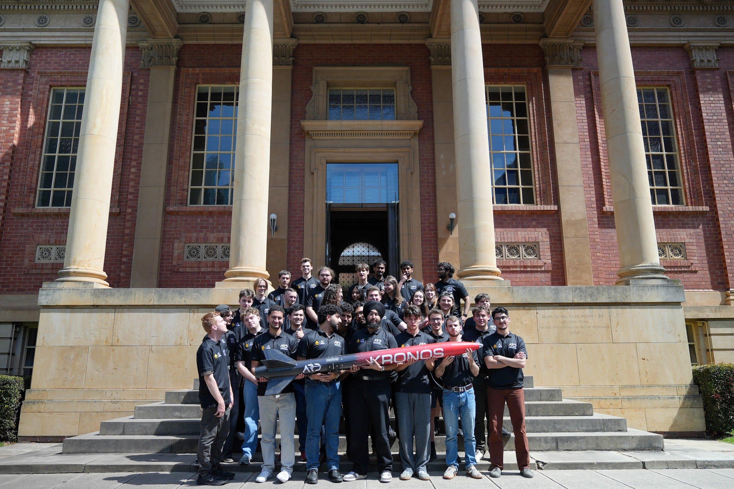A large group of team members in black ARD shirts stands on the steps of a sandstone and red-brick building, with the front row holding the Kronos rocket horizontally.