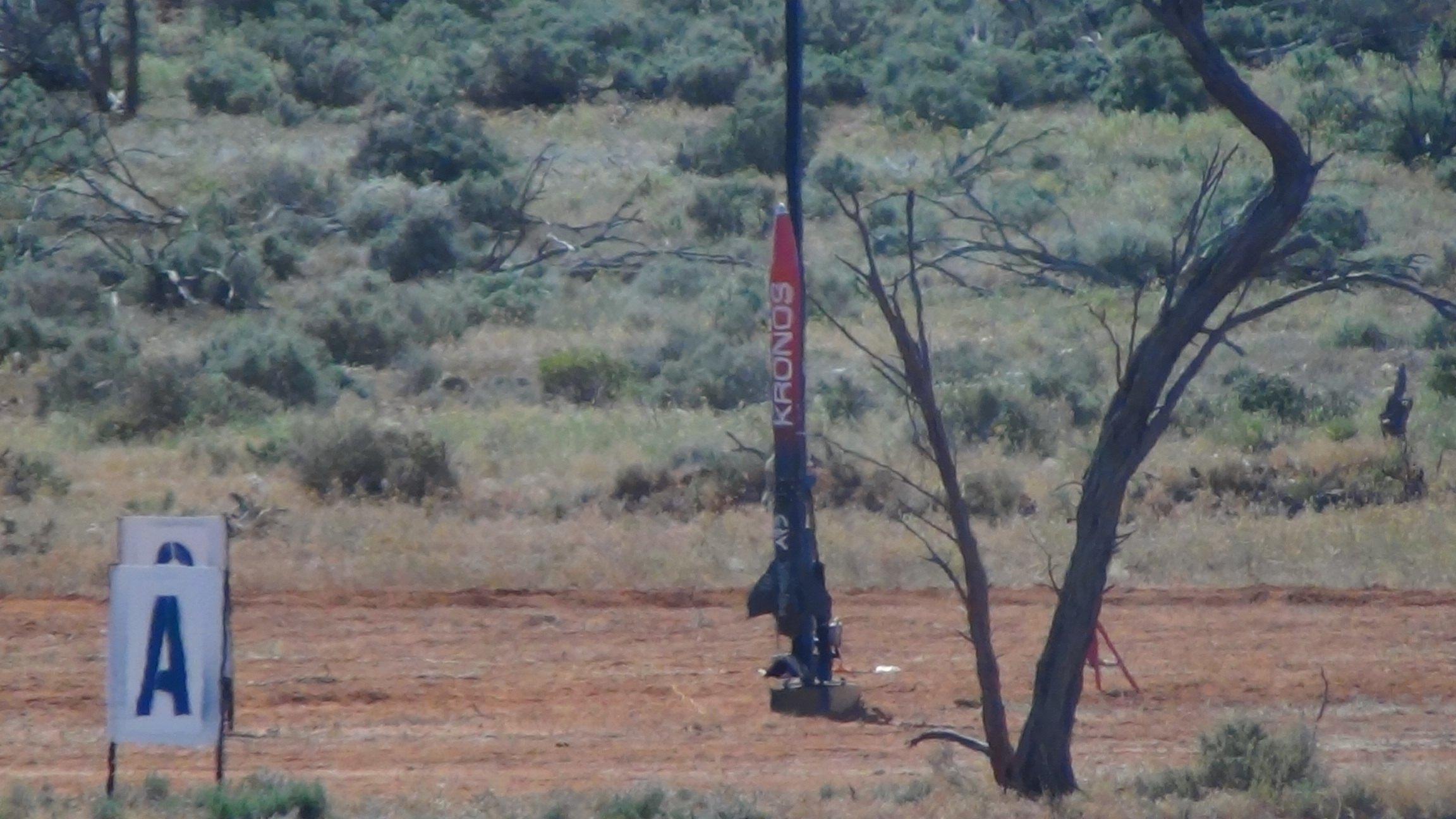 Kronos stands vertically on the launch rail beside a small tree in scrubland, with a pad marker sign reading "A" in the foreground.