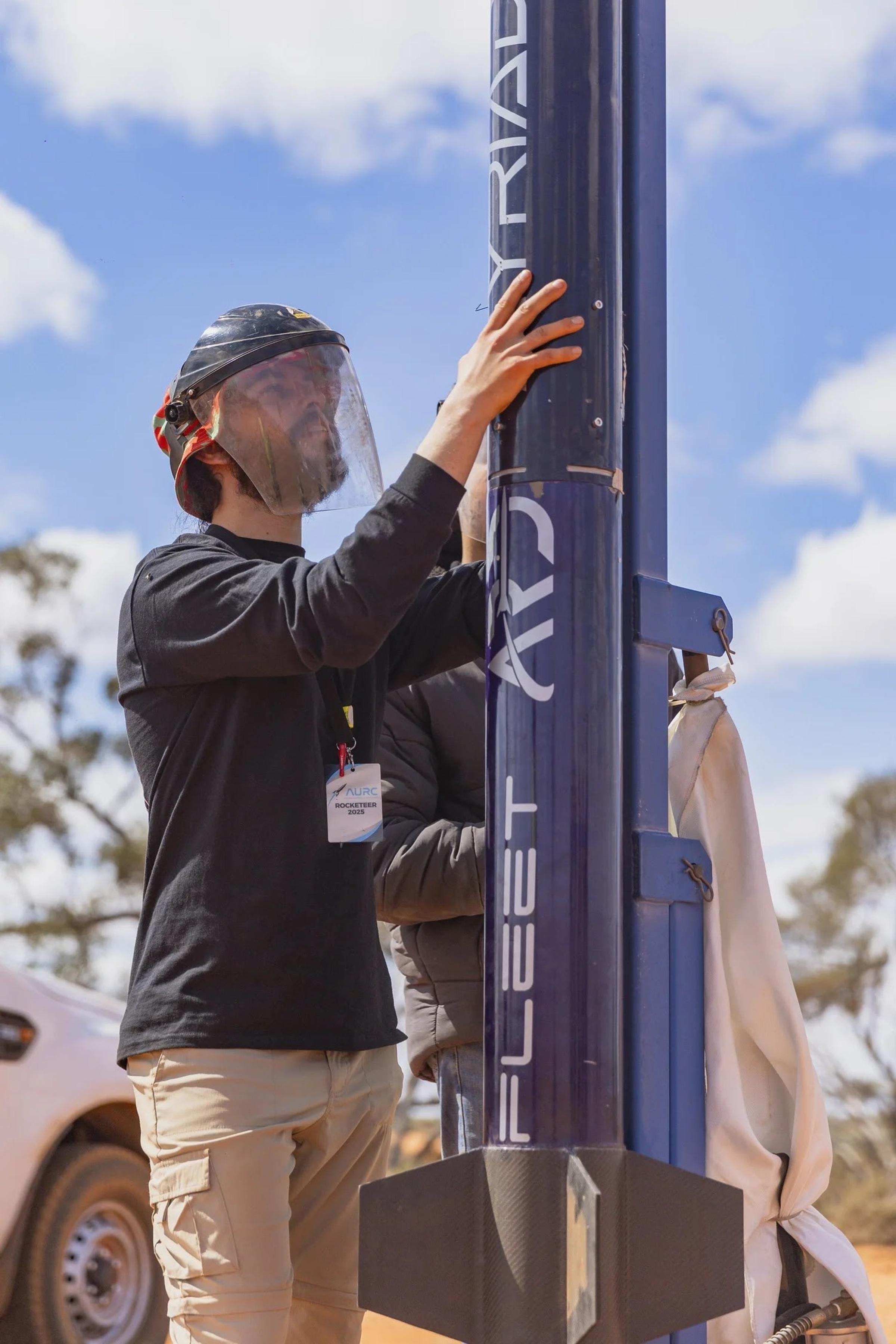 A team member in a face shield stands beside the navy Myriad rocket mounted on a blue launch rail, with "MYRIAD" lettering running up the body.