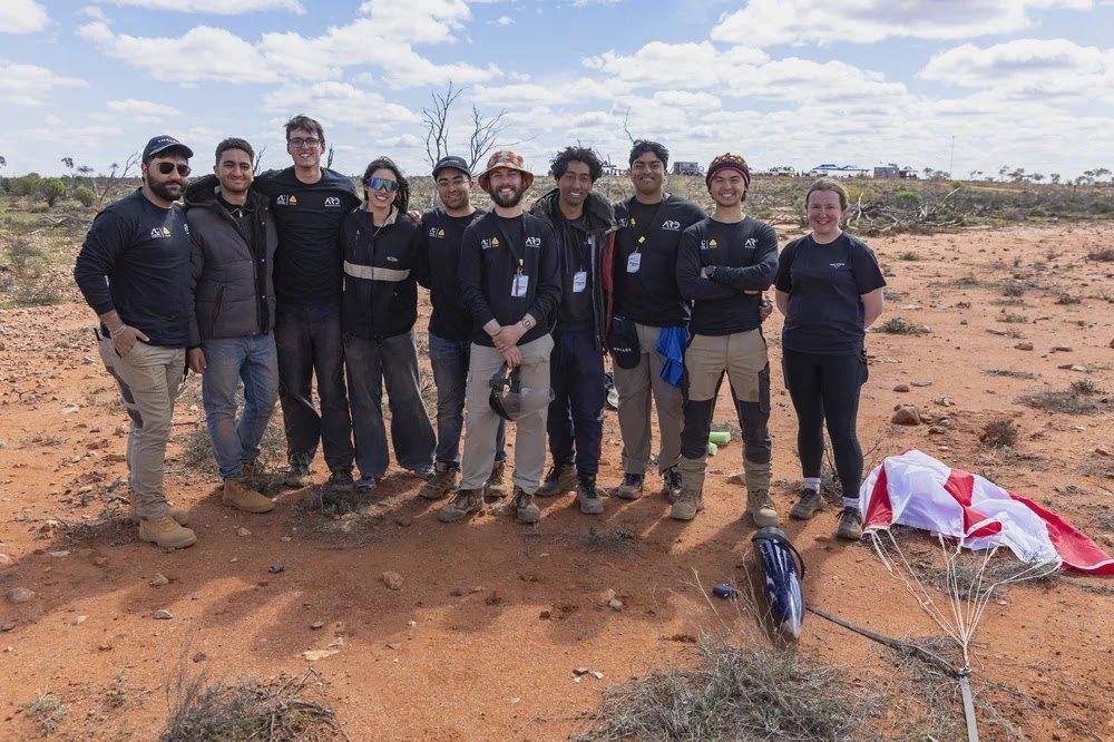 A group of team members stands behind the recovered Myriad rocket and its red and white parachute on red desert ground.