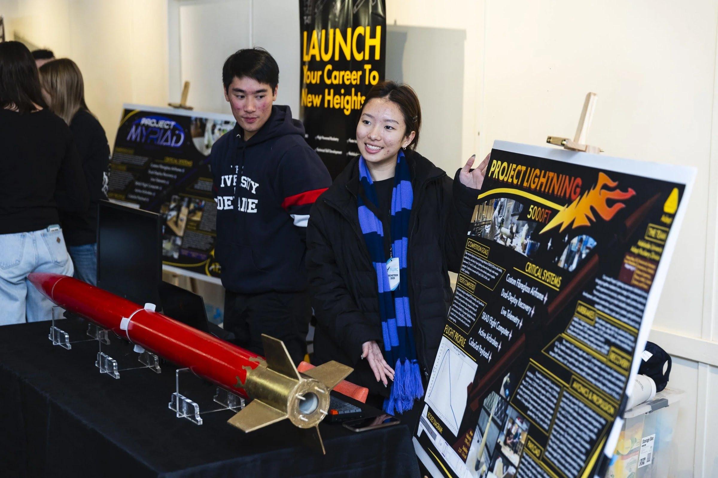 Two team members stand behind the red Lightning rocket with gold fins on clear display stands, beside "Project Lightning" and "Project Myriad" posters.