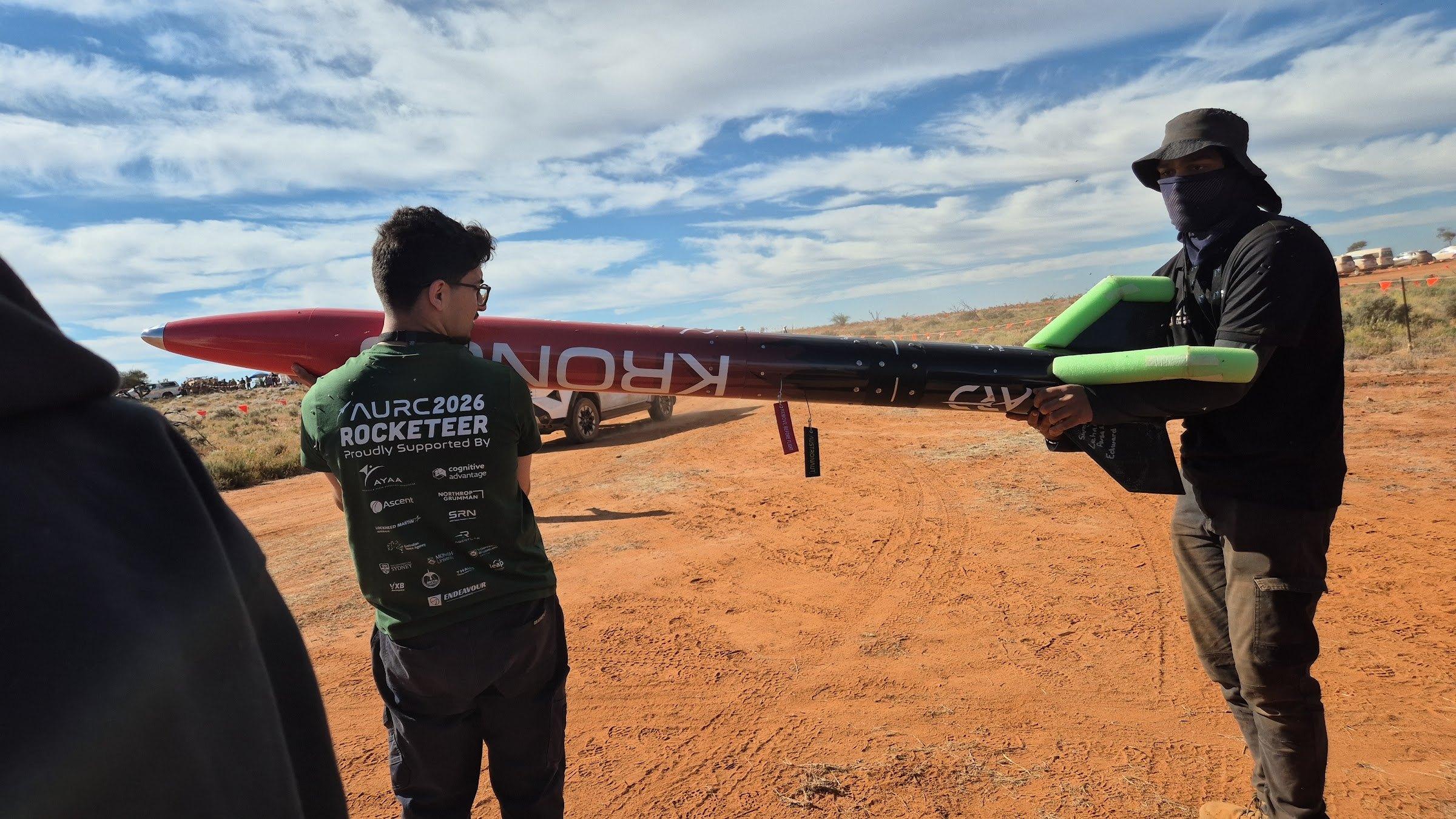 Two team members carry the assembled Kronos rocket horizontally across red dirt under a blue sky with scattered clouds.