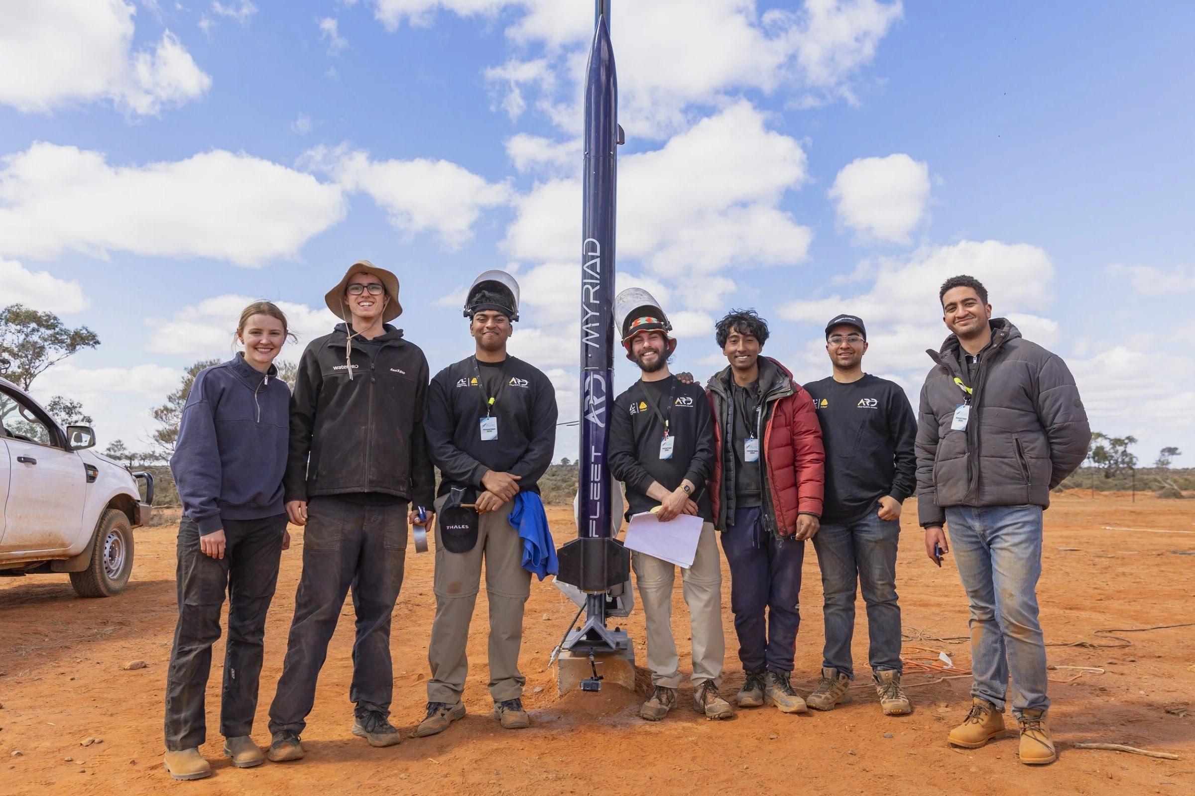 A line of team members stands on red dirt in front of the Myriad rocket mounted vertically on its launch rail.