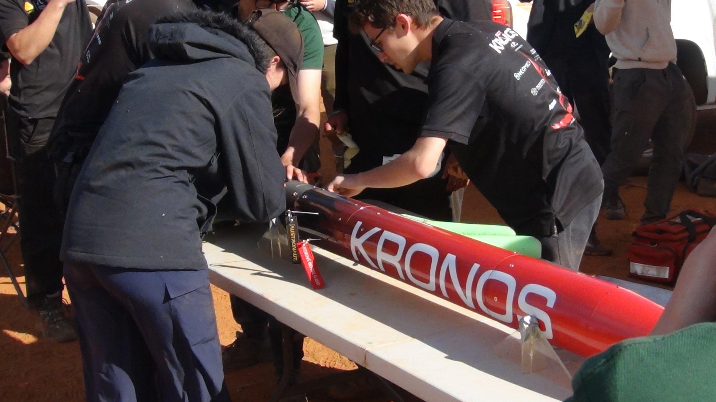 Under a shade tent, team members lean over a long table to assemble the red Kronos rocket, its white KRONOS lettering visible along the body.