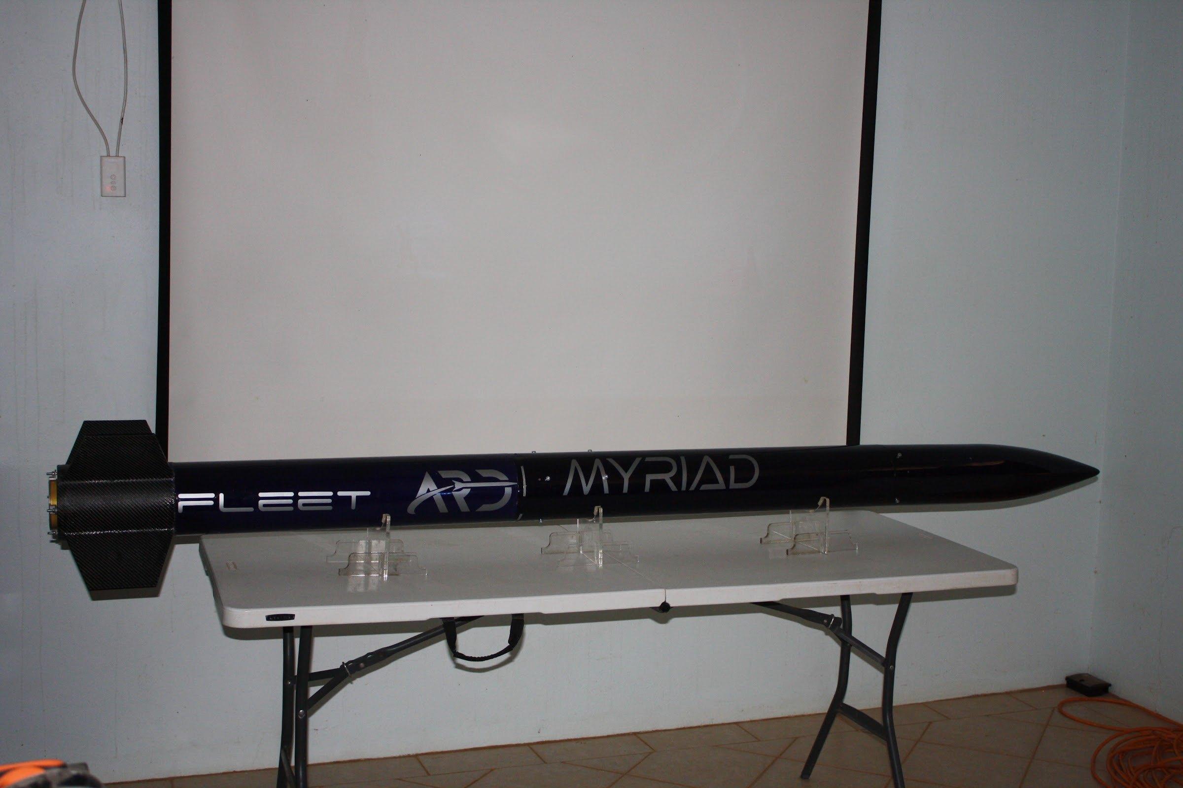 Side view of the Myriad rocket resting on clear stands on a white table: a dark navy body with "FLEET", an ARD logo and "MYRIAD" in white, a black ogive nose cone and a black finned tail.