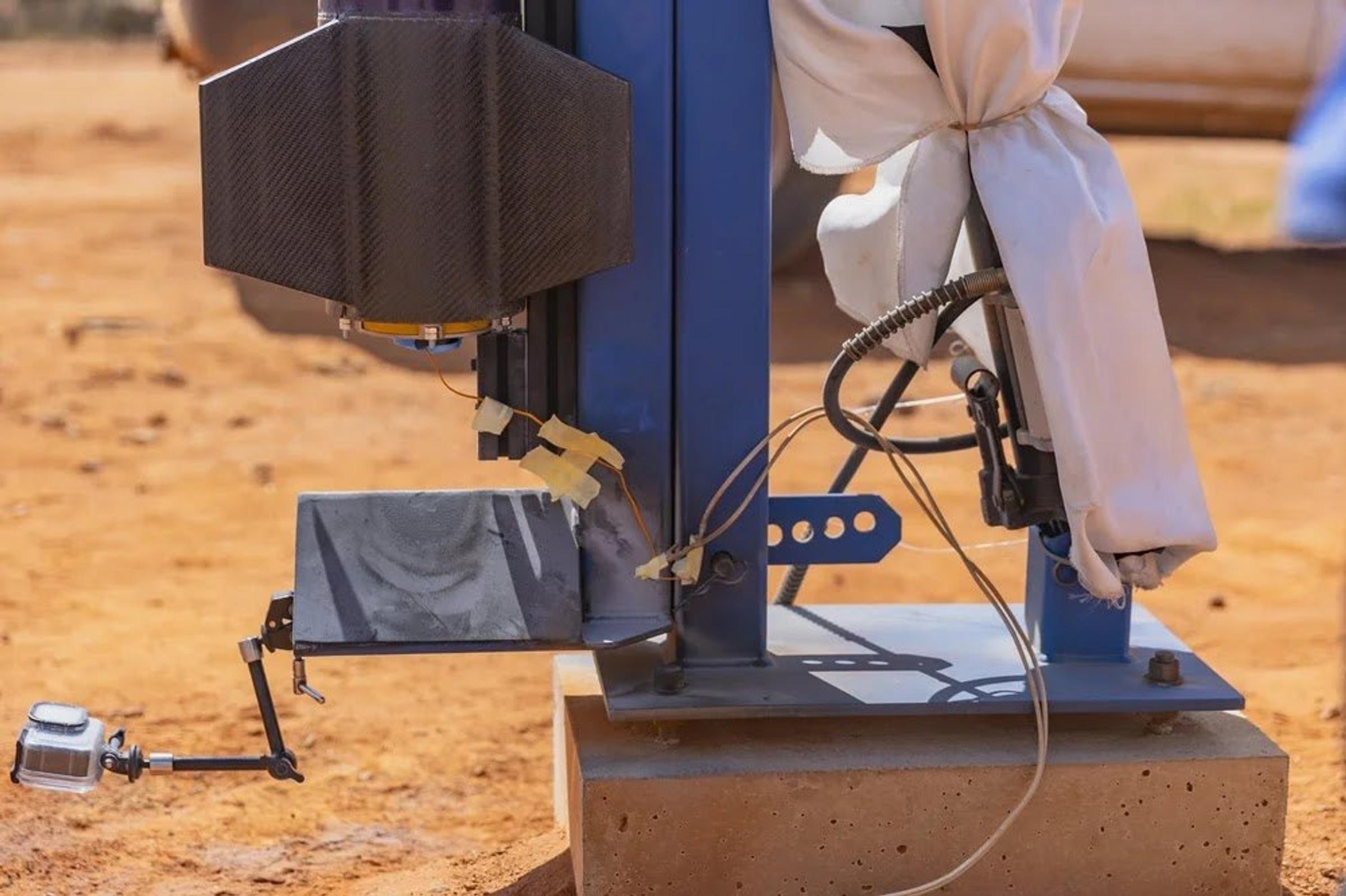Close-up of the base of a blue launch rail with a steel blast deflector, ignition wiring and a white cloth-wrapped fitting on a concrete footing.