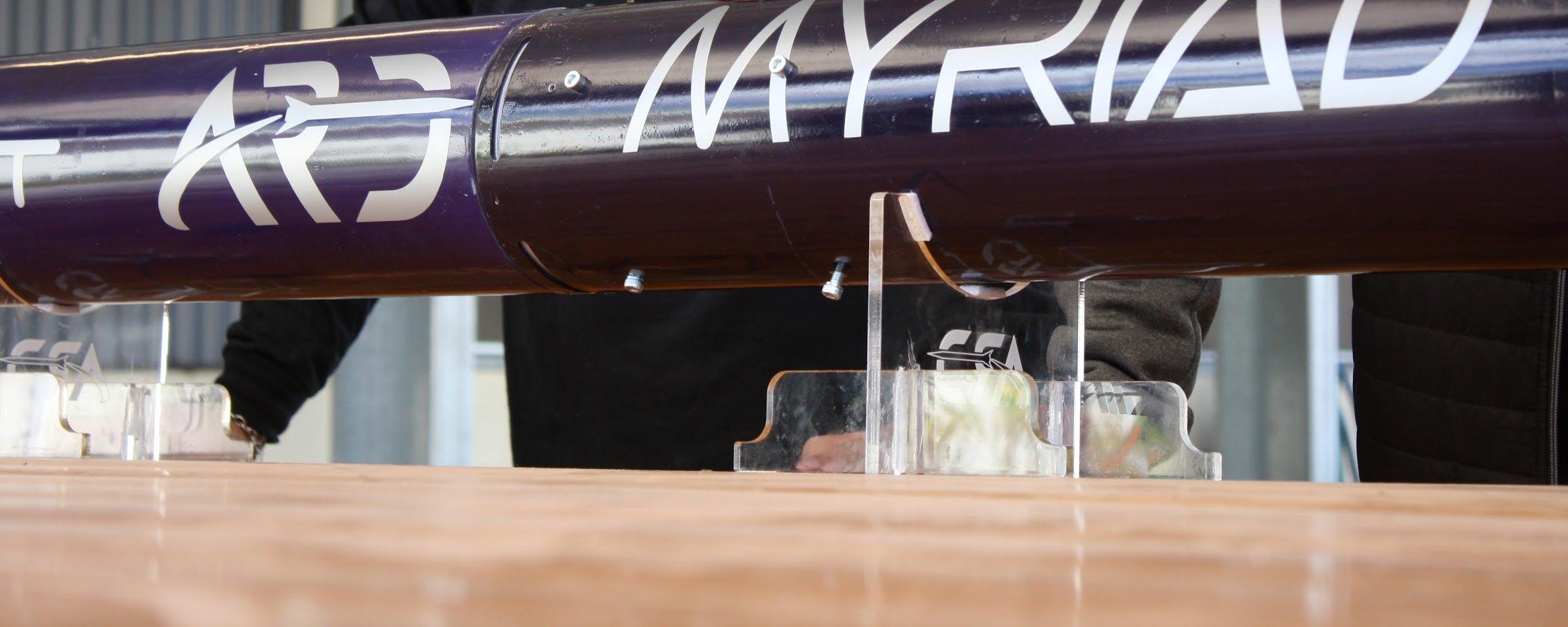 Close-up of Myriad's glossy navy airframe on clear acrylic stands, showing the white ARD logo and "MYRIAD" lettering across a body-tube joint.