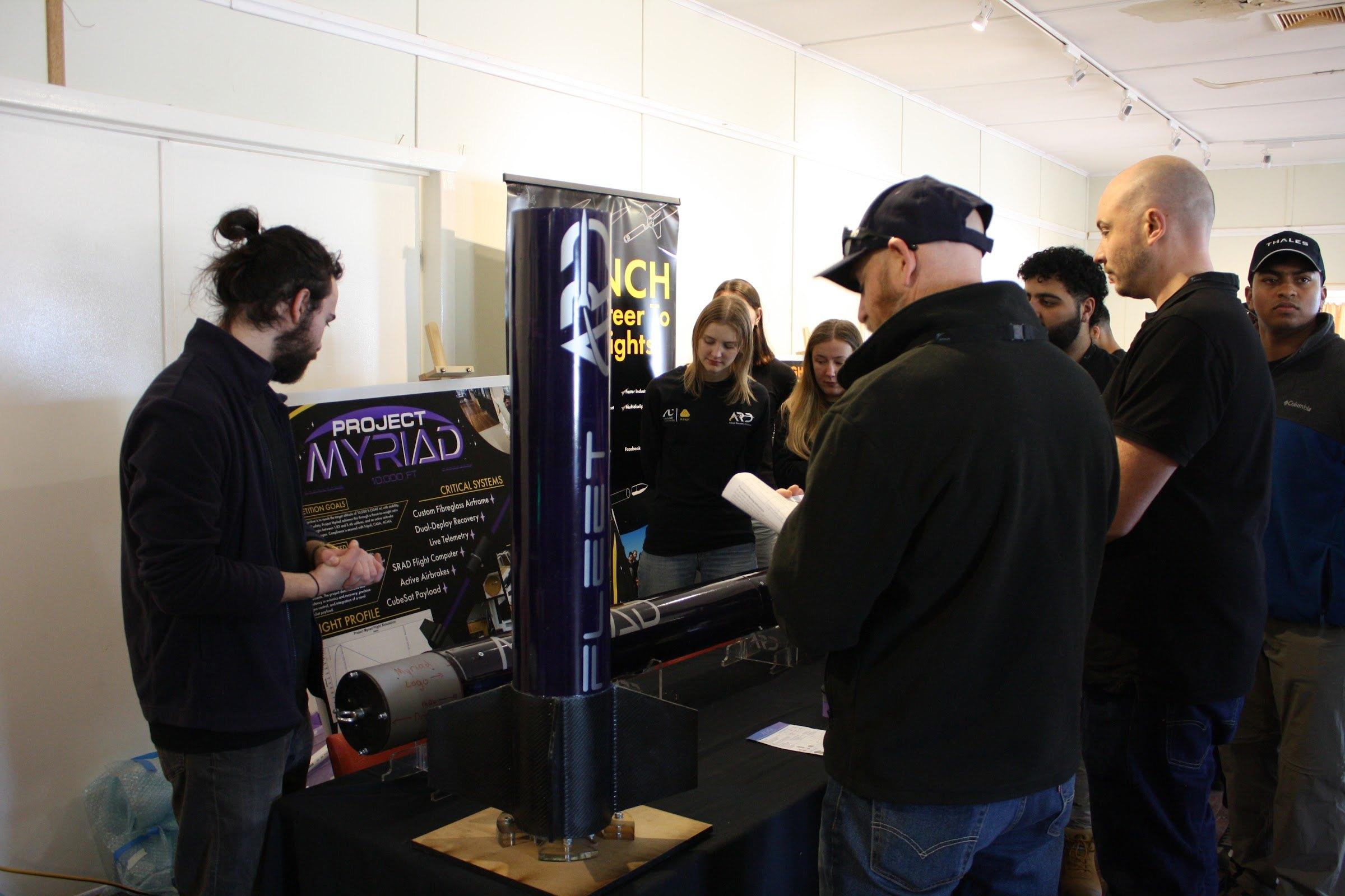 Myriad's upper section stands upright on a display table beside a "Project Myriad" poster while visitors talk with team members.