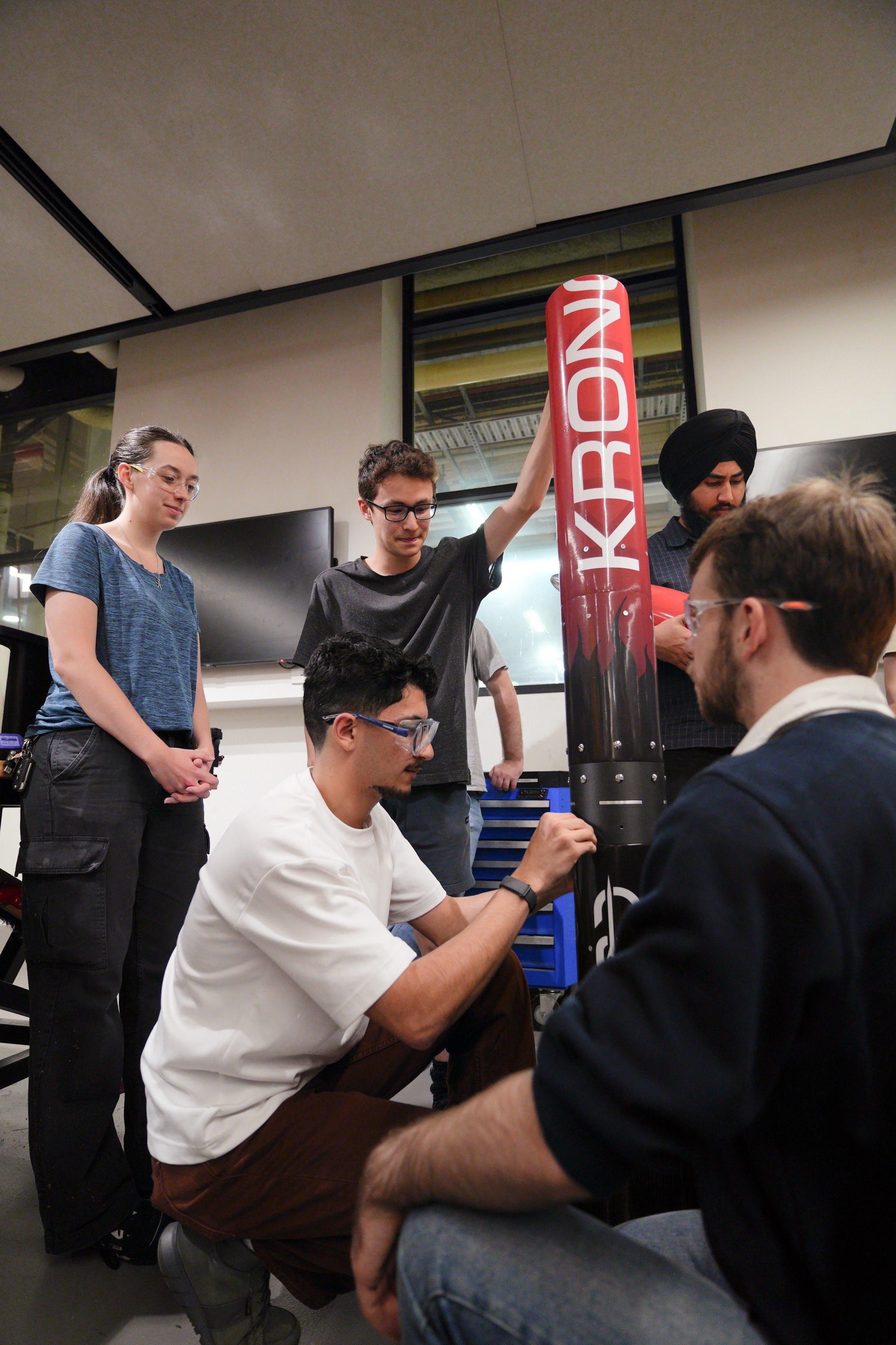 In a workshop, team members gather around the upright Kronos rocket while two of them kneel to work on its lower body section.