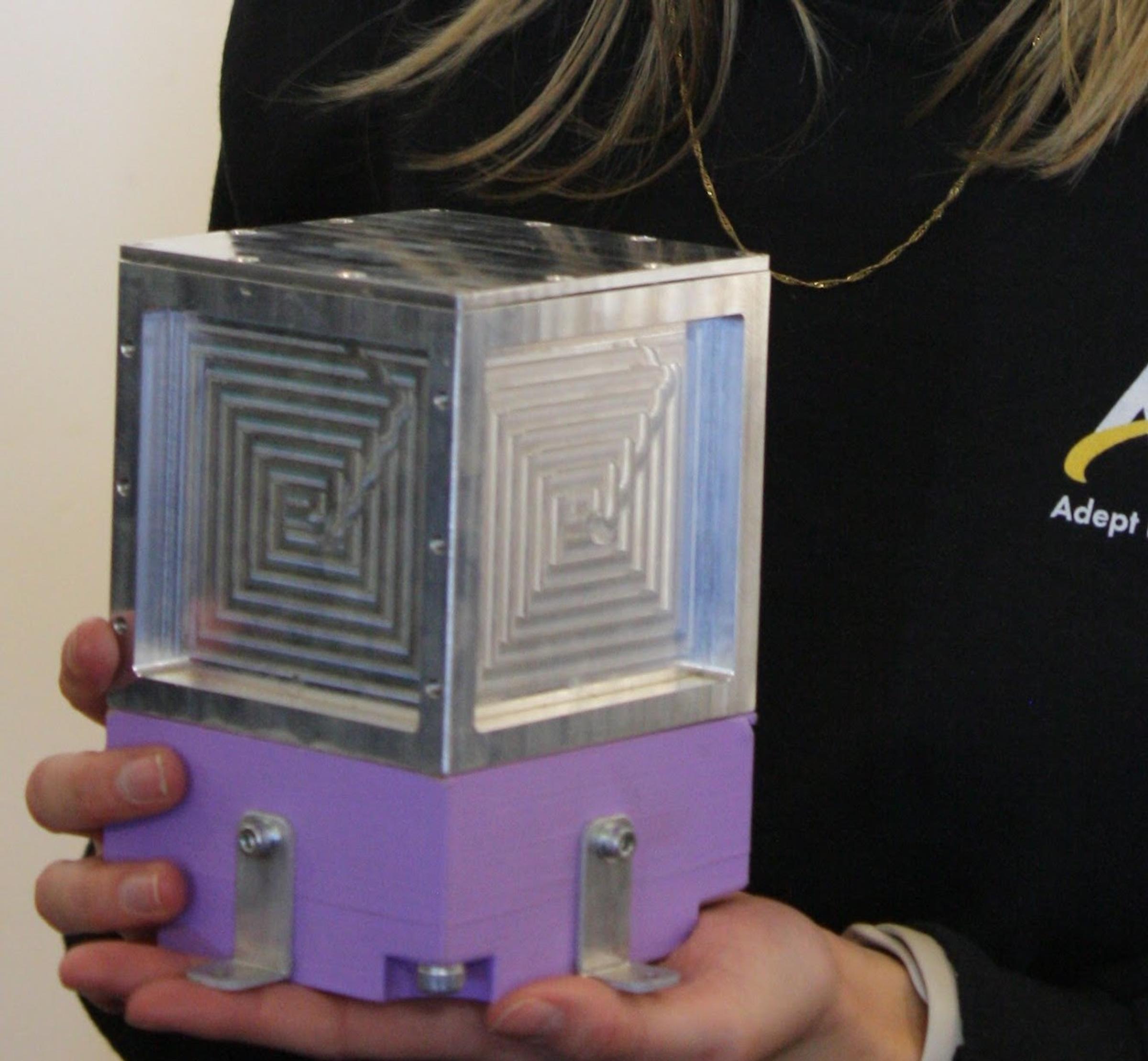 A machined aluminium cube with stepped square patterns on its faces, mounted on a purple 3D-printed base with metal brackets, held in two hands.
