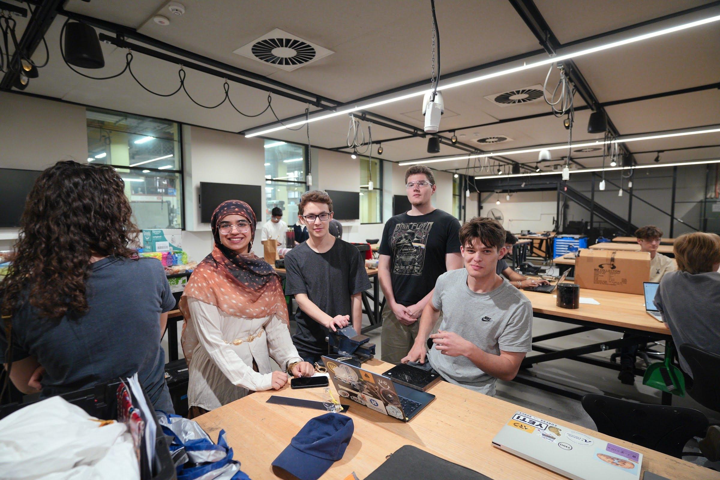 Team members sit and stand around a wooden workbench with laptops in a large, brightly lit workshop.