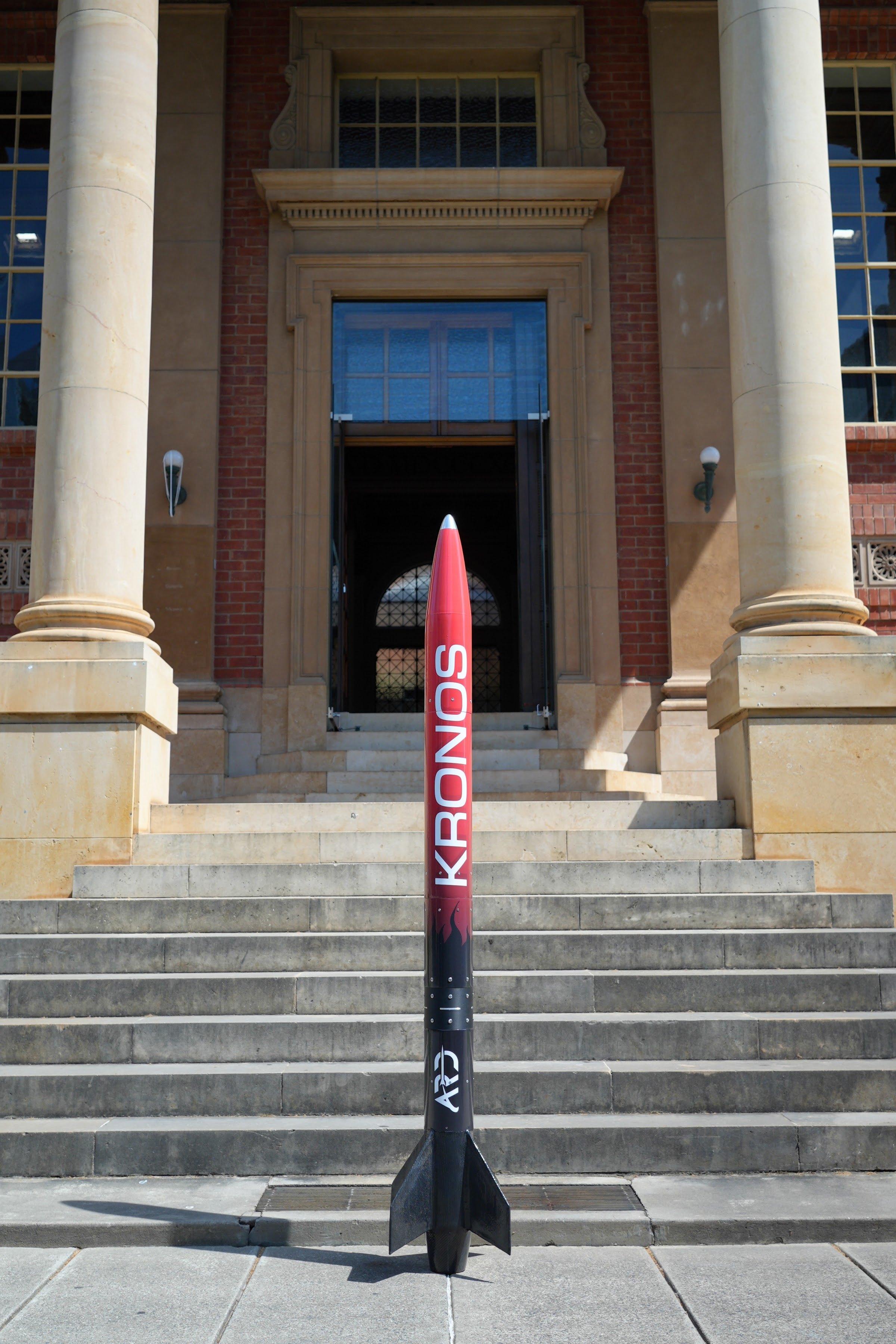 The Kronos rocket standing upright on its fins at the foot of a stone staircase between two columns; red nose cone, red-to-black fade along the body, vertical white KRONOS lettering and an ARD logo near the black finned tail.