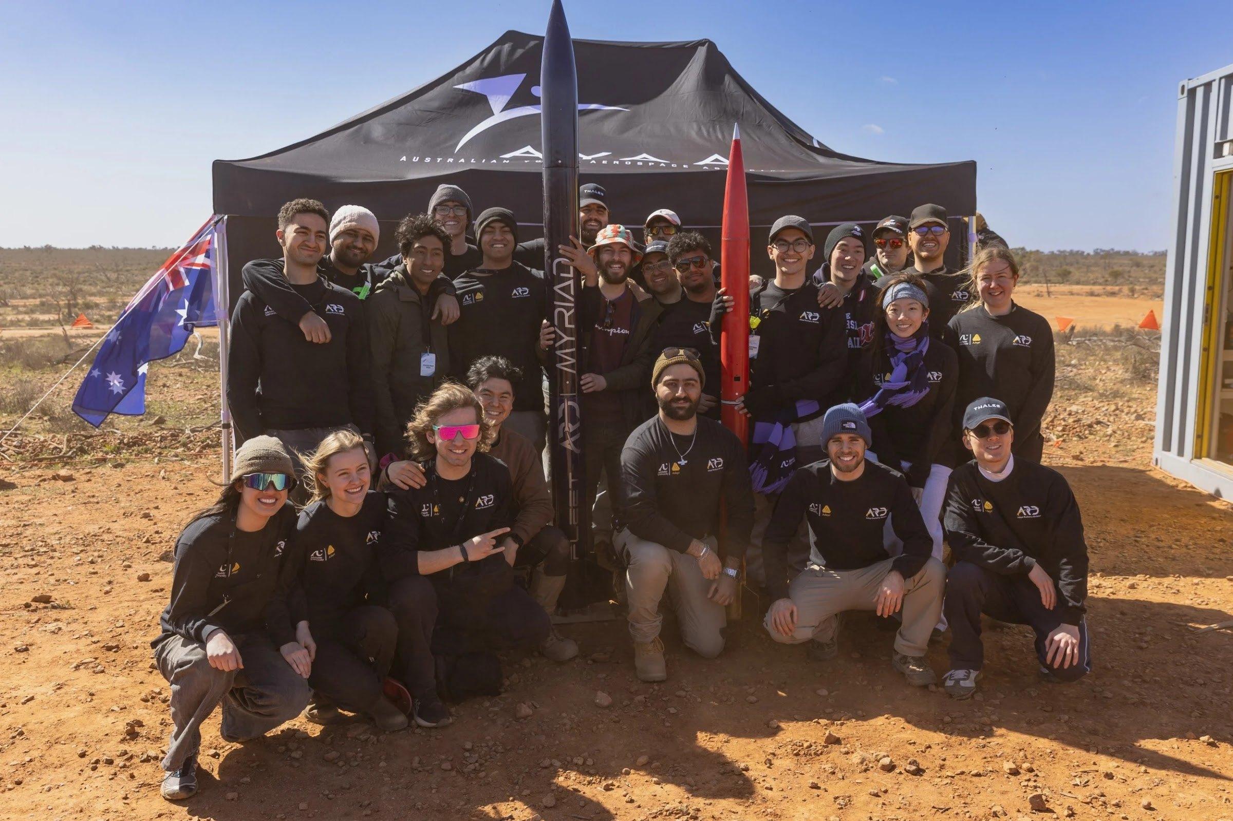 A large group of team members poses in front of a black shade tent on red dirt, with an Australian flag and two rockets standing upright among them, one navy and one red.