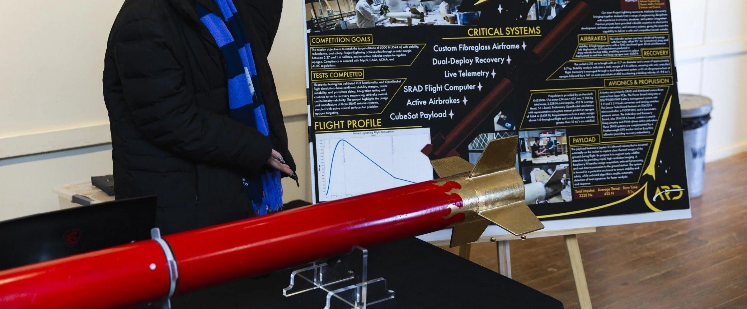 The red Lightning rocket with a gold fin can and gold fins lies on a display table in front of a black "Project Lightning" poster.