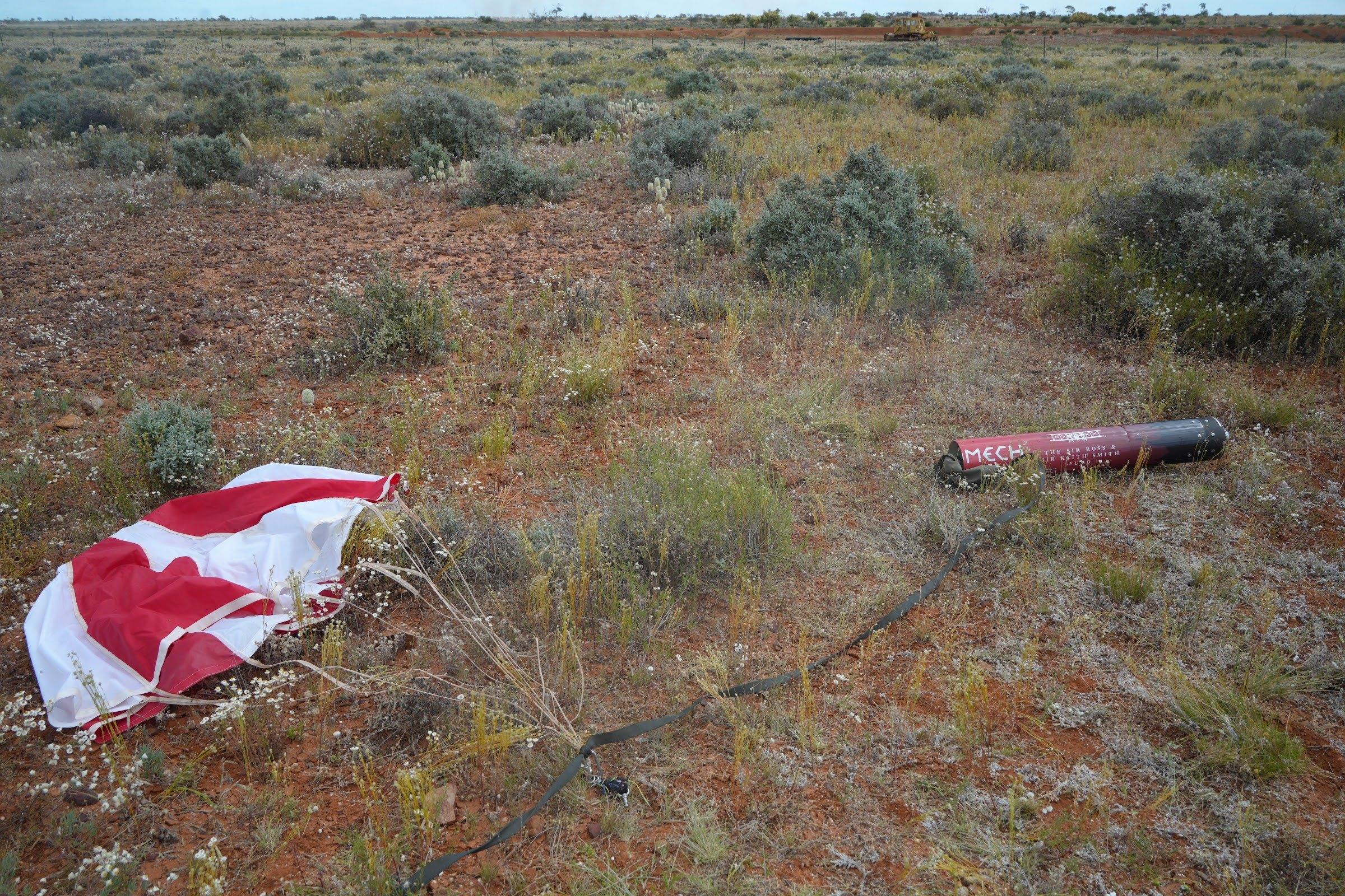 A red and white parachute lies spread over low scrub, with a red rocket body section on the ground further back.