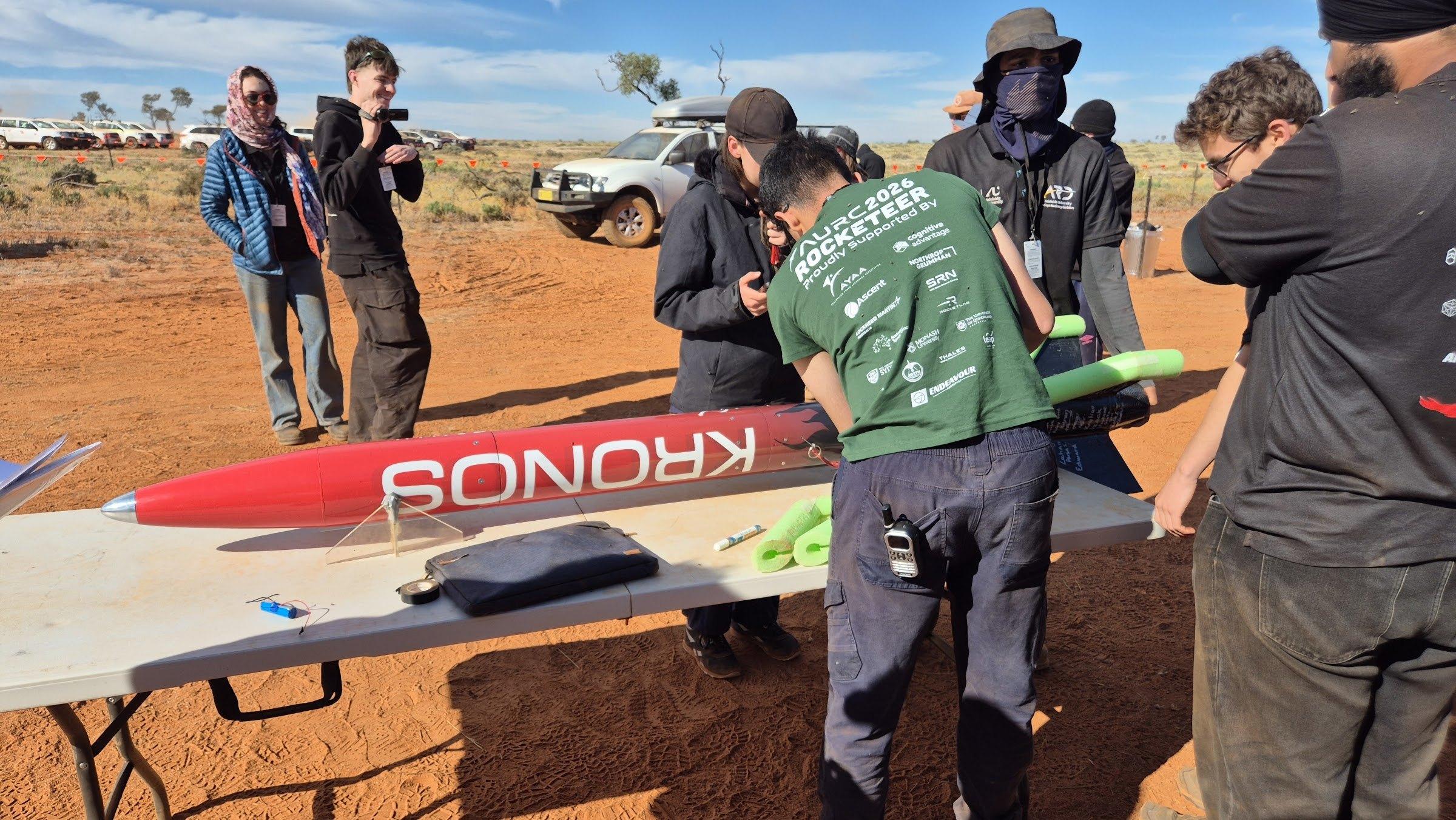 Kronos lies on a folding table on red desert ground while team members and officials in hi-vis gather around it.