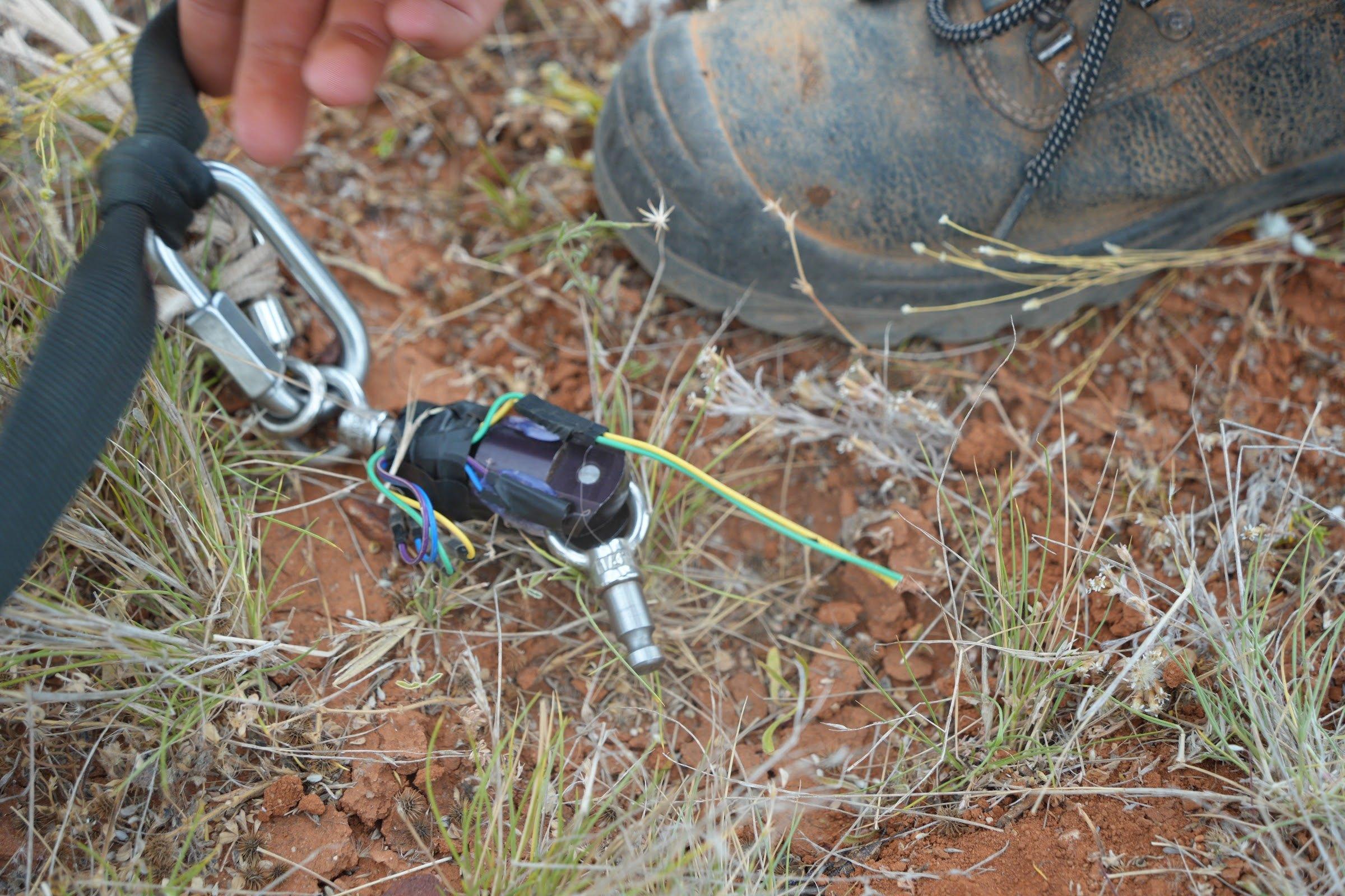 Close-up of black recovery webbing joined by a steel quick link and swivel to a small black electronic unit with coloured wires, lying in dry grass.