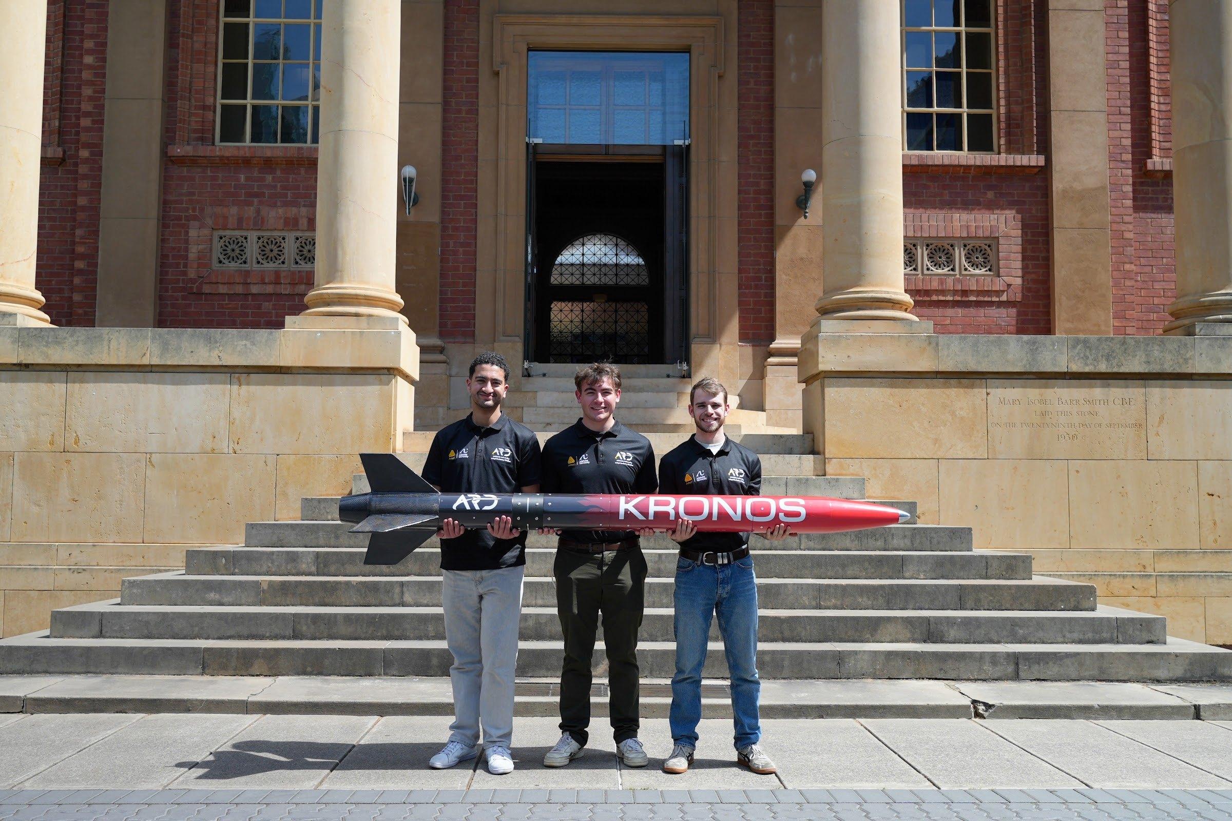 Three team members in black polo shirts hold the Kronos rocket horizontally in front of a sandstone and red-brick building; the rocket is black at the finned tail and fades to red towards the nose, with white "KRONOS" lettering.