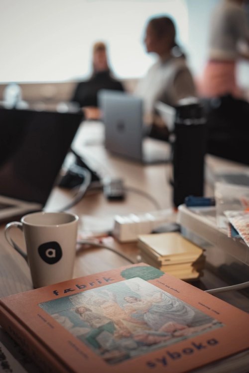 A Fæbrik sewing book and an Agens mug on a desk, with colleagues working behind.