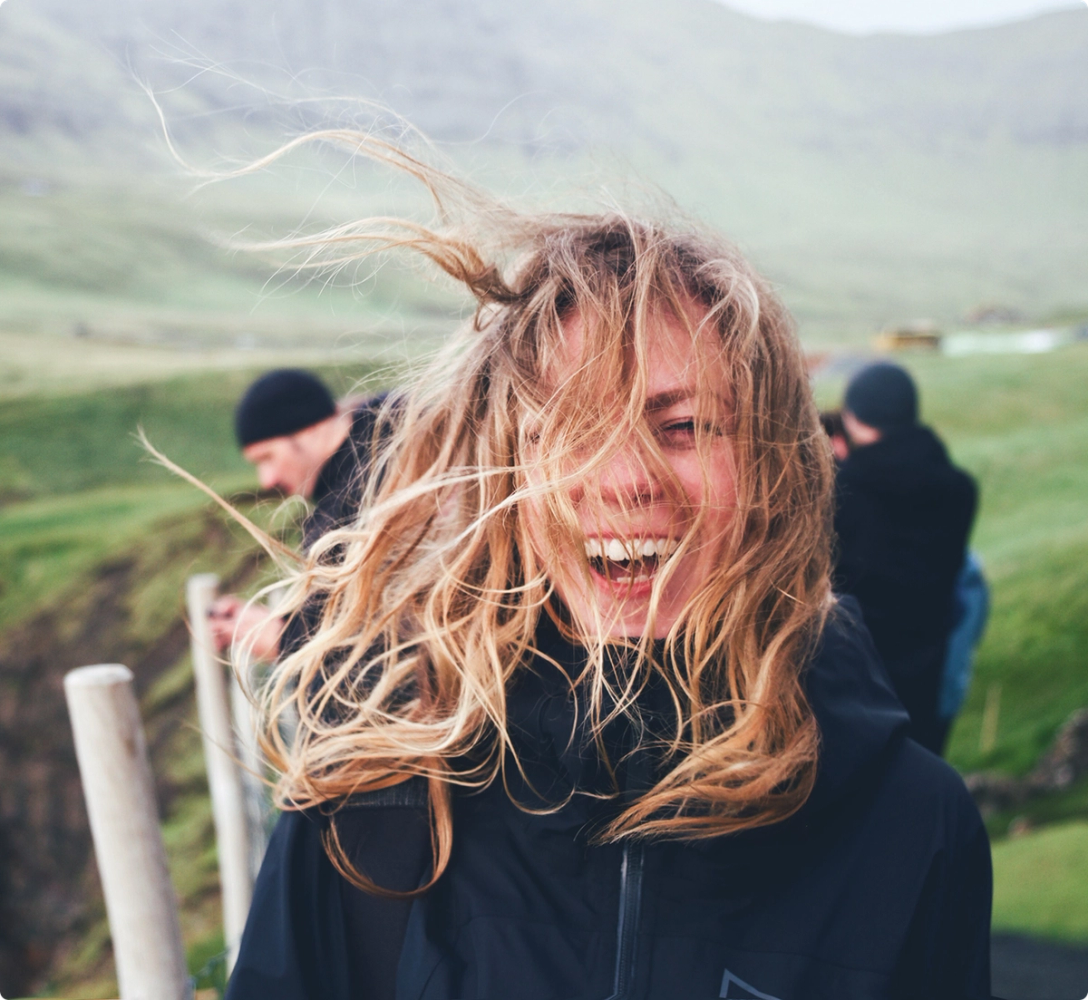 A colleague laughing into the wind on a green coastal hillside, hair blown across their face.