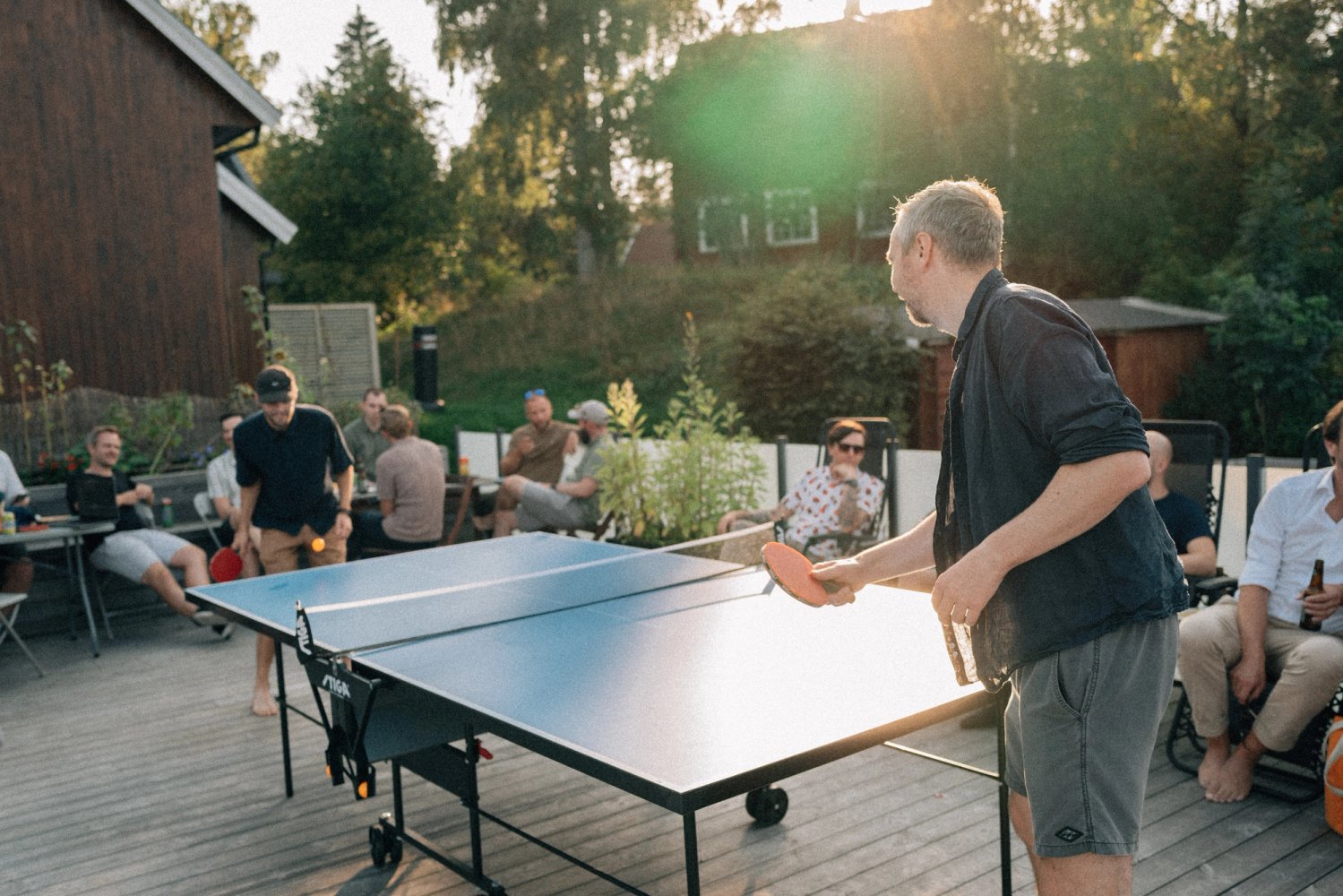 A table-tennis rally on the office deck in evening sun, colleagues watching from the side.