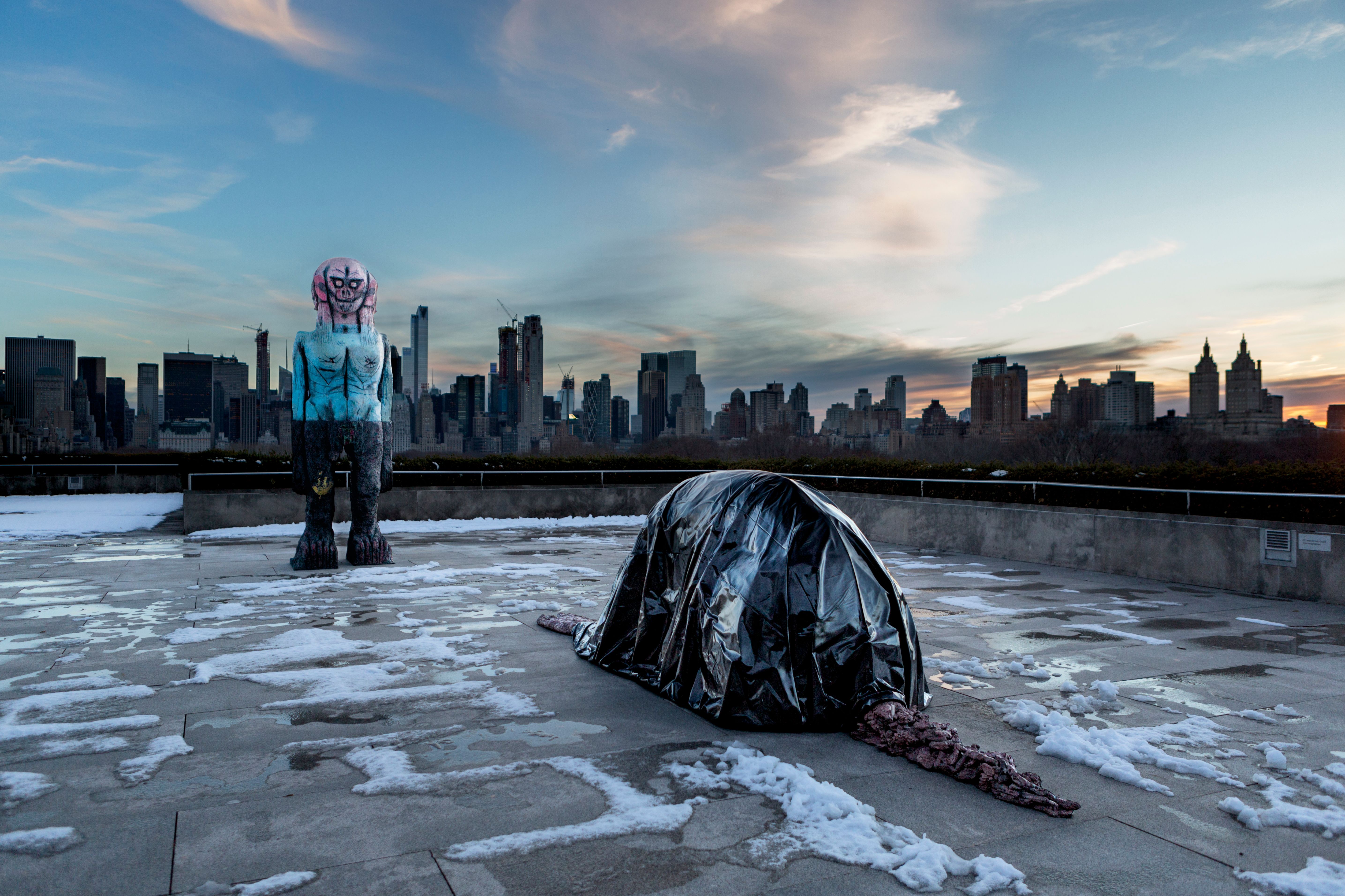 On a rooftop of the MET in New York City, we see two sculptures against the skyline—one of a totem figure looking toward the camera, and another of a figure kneeling away from the camera.