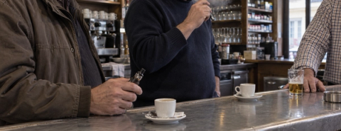 Three men stand at a bar counter with coffee cups and a glass of beer; one holds a small vaping device.