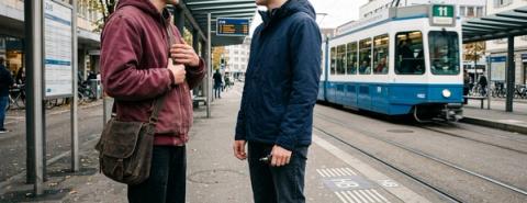 Deux jeunes hommes discutent à un arrêt de tramway, à côté d’un tram bleu et blanc.