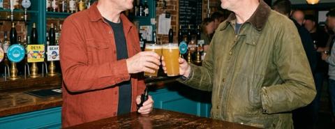 Two men holding pints of beer at a pub bar, one also holding a vape.