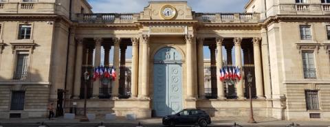 Grand neoclassical stone building with tall columns, French flags flanking a large blue door, and a black car parked outside.