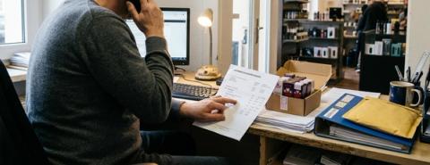 A vape shop worker sits at a desk holding a supplier invoice beside a box of e-liquid products, with the shop visible in the background.