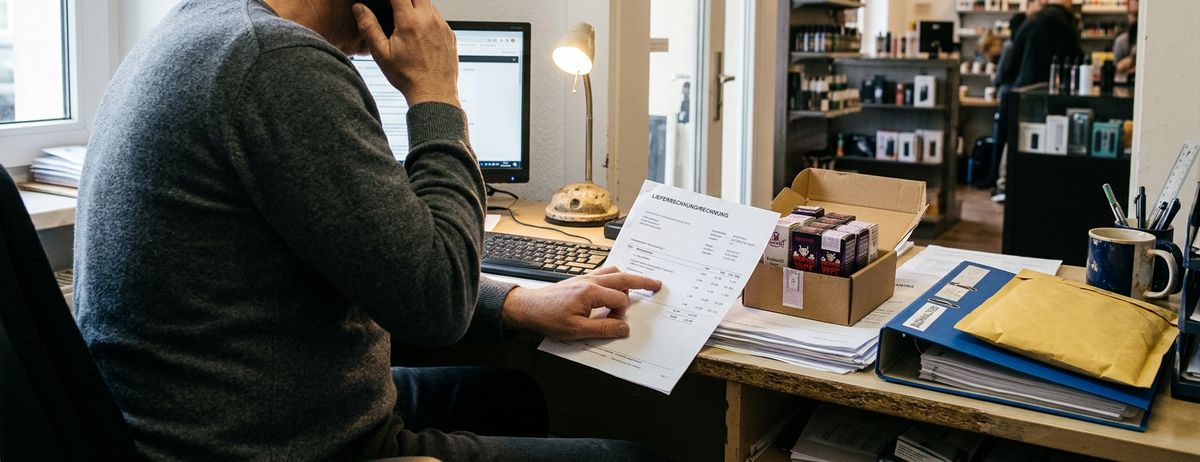 A vape shop worker sits at a desk holding a supplier invoice beside a box of e-liquid products, with the shop visible in the background.
