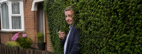 Illustration of a man holding a cigarette beside a hedge outside a brick house.