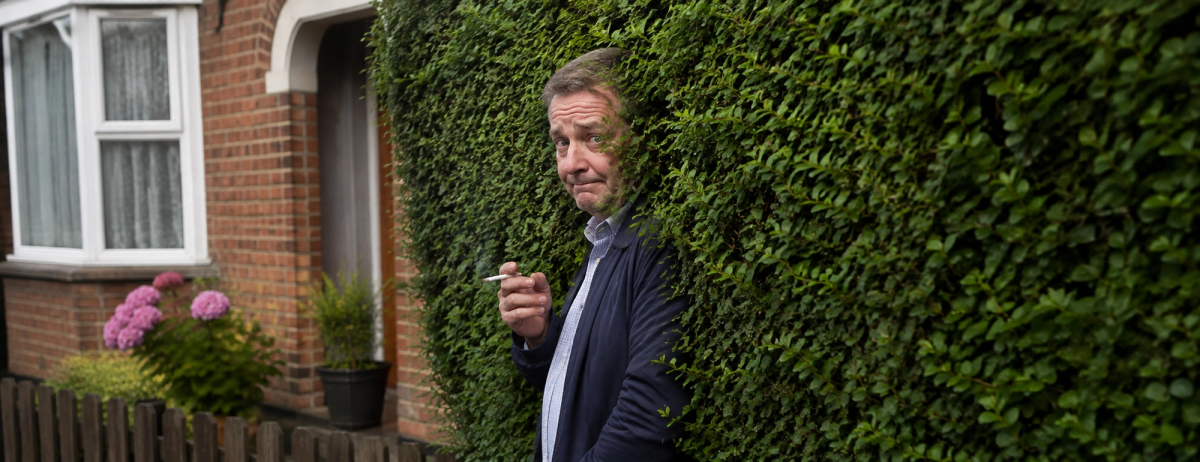 Illustration of a man holding a cigarette beside a hedge outside a brick house.