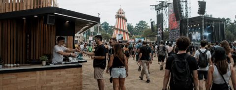 Festival-goers beside a wooden stall displaying round product tins, with a concert stage in the background.