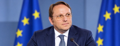 A man in glasses sits at a microphone with European Union flags behind him.