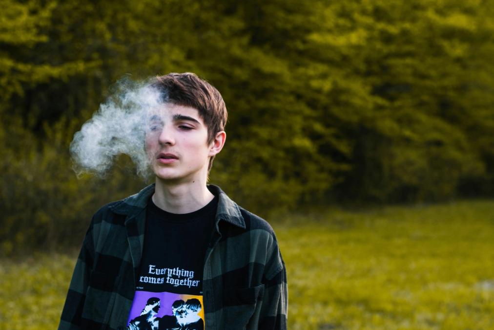 A person exhales a cloud outdoors against a background of green trees.