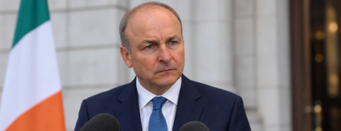 A man in a suit stands behind microphones beside an Irish flag.