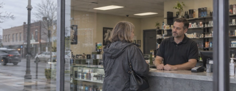 A customer speaks with a man behind the counter of a vape shop, with shelves of boxed products and bottles behind him.